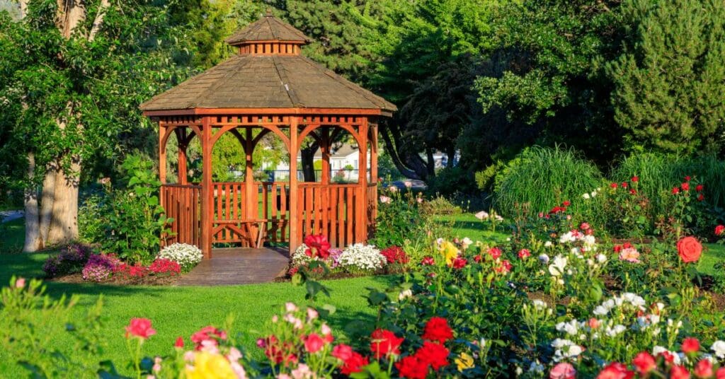 An outdoor wooden gazebo placed in a landscaped yard with greenery and grass.