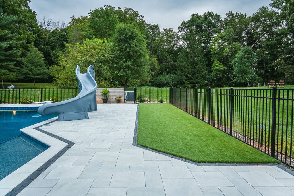 Modern backyard with a swimming pool, curved water slide, stone paver patio, artificial grass strip, and black metal fence surrounded by lush trees